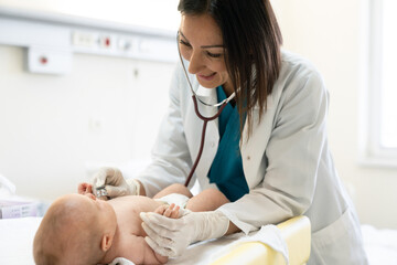 smiling female pediatrician using stethoscope and protective gloves to check up a newborn baby.