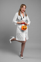 Wall Mural - Happy woman in scary nurse costume with pumpkin bucket on light grey background. Halloween celebration