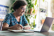 © PEDROMERINO - Old woman with glasses using a laptop and a notebook at home