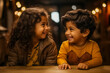© fotogurmespb - Two joyful children exchange smiles across a table in a warm, bokeh-lit setting, capturing a moment of childhood happiness and innocence