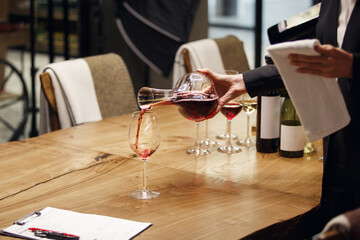 Professional female sommelier pours red wine from decanter to the glass, close up image. Woman waiter pouring alcoholic drink being in cellar of wine shop.