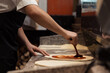 © arthurhidden - A chef in a professional kitchen applies tomato sauce evenly on stretched pizza dough, showcasing pizza preparation