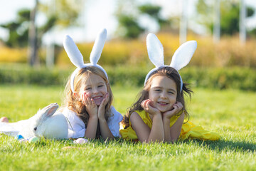 Sticker - Two little girls with a bunny rabbit have an Easter at green grass background. Two children wearing Bunny ears find and pick up multicolored egg on Easter egg hunt in garden.