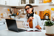 © dvulikaia - Portrait of happy female student watching webinar or tutorial studying online on laptop sitting at kitchen table, noting down after teacher or lecturer.