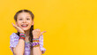 A little girl points her finger at your advertisement. The child put on a lot of beaded bracelets. Beading for children. Yellow isolated background. Copy the space.