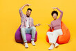 Full body smiling satisfied young couple two friends family man woman wear purple casual clothes together sit in bag chair point index finger on area between them isolated on plain yellow background.