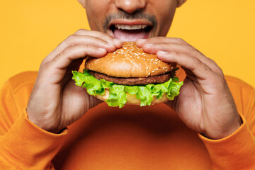 Wall Mural - Close up cropped young man wear orange sweatshirt casual clothes holding eating biting burger isolated on plain yellow background studio. Proper nutrition healthy fast food unhealthy choice concept.