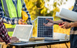 © NanSan - engineers discussing a solar energy project with a small solar panel model on the table, a laptop, blueprints, and a wind turbine model, likely for a clean energy city initiative.