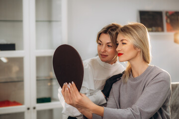 Wall Mural - Bottom view of mirror reflection of smiling caucasian girl looking at her beautiful lips while female beautician looking at her after permanent makeup lips procedure in beauty salon.