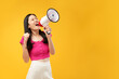 Photo of a young Asian woman holding a megaphone and shouting aggressively with fists clenched. Wearing a pink t-shirt, and isolated on a yellow background.