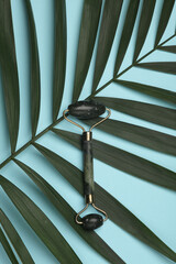 Wall Mural - Jade massage roller with green palm leaf on blue background. Creative beauty layout