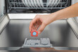 Young adult woman hand fingers holding and adding blue red dishwasher tablet in detergent dispenser for dishes washing. Closeup. Front view.