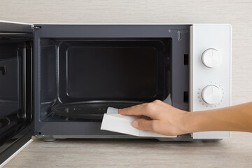 Young adult woman hand holding dry white paper napkin and wiping dark black surface of white microwave oven inside on table top at home kitchen. Closeup. Regular cleanup. Front view.