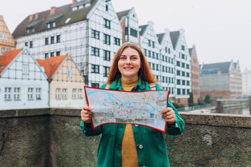 Wall Mural - Attractive young female tourist is exploring new city. Redhead girl with backpack holding a paper map on city street in Gdansk. Traveling Europe in autumn.