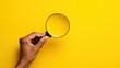 a person's hand holding a magnifying glass over a yellow background with copy space in the middle.