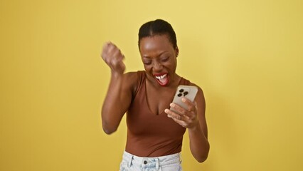 Wall Mural - Excited african american woman, proudly celebrating victory and success, raises arm whilst texting on her yellow phone against an isolated background