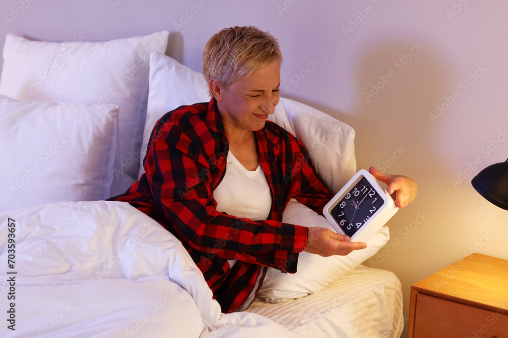 Displeased mature woman with clock in bed at night
