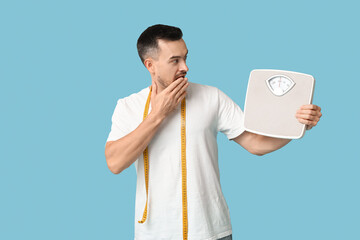 Wall Mural - Handsome sporty shocked man with measuring tape and scales on blue background. Weight loss concept