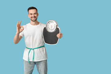 Wall Mural - Handsome sporty young man with measuring tape and scales showing OK gesture on blue background. Weight loss concept