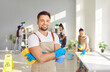 Portrait of happy smiling male young janitor in uniform looking cheerful at camera holding household cleaning supplies in hands. Cleaning team service, housework and housekeeping concept.