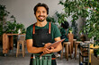 Handsome confident sincere smiling male gardener florist in green t-shirt black apron and bag of garden tools on belt holding wooden clipboard and pen looking at camera. Work in a plant shop.