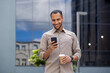 © Liubomir - Businessman walking on break outside office building, male holding phone in hands, using app on smartphone to browse social network, holding cup of hot coffee drink in hand, smiling joyfully.