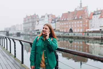 Wall Mural - Young happy redhead woman making phone call over mobile phone in the city. Middle age woman talking on smartphone on urban background. Traveling Europe in autumn