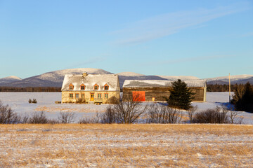 Wall Mural - Golden morning winter view of steep roofed patrimonial fieldstone house and wooden barn with the Laurentian mountains in the background, St. François, Island of Orleans, Quebec