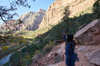 A tourist explores a trail in zion national park with her phone camera. She snaps a picture of the virgin river and nearby mountain peaks.