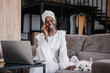 Smiling African woman in a headscarf having a pleasant conversation on a mobile phone stroking dog at home. Girl in turban enjoying phone talk with friend.