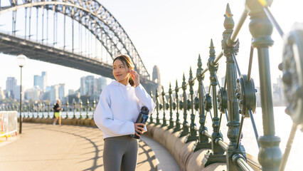 Wall Mural - Happy Asian woman listening to music from mobile phone and earphones with mobile app during jogging exercise at city street in the morning. Healthy girl do sport training running workout in the city.