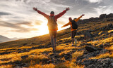 Fototapeta Natura - Couple hikers are walking together in sunset mountains. Two young tourists with backpacks are standing with open arms and enjoys sunset