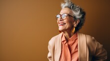  A Woman With Grey Hair Wearing Glasses And A Pink Shirt And A Tan Blazer Smiles At The Camera With Her Hands Behind Her Back To The Camera.