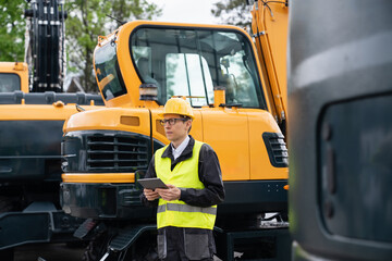 Wall Mural - Engineer in a helmet with a digital tablet stands next to construction excavators.