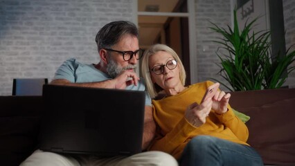 Poster - Senior couple using a smartphone and a laptop while seated at the sofa in the living room at home. A mature couple in love sitting in the living room in a cozy home, using a laptop and phone