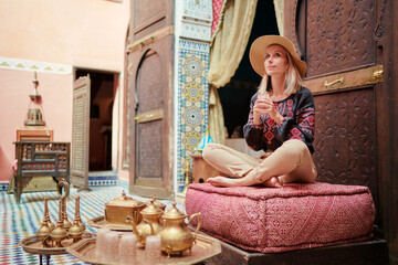 Wall Mural - Traveling by Morocco. Happy young woman in hat relaxing in traditional riad interior in medina.