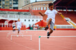 Black little boy jumping over hurdles on running track at stadium.
