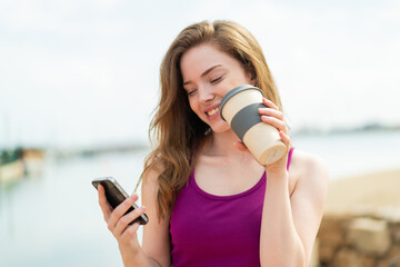 Canvas Print - Young redhead woman at outdoors using mobile phone and holding a coffee with happy expression