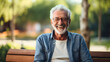 Happy old aged senior man or grandpa with gray hair and beard, sitting on the wooden bench in nature park, wearing glasses, smiling and looking at the camera. Male pensioner person leisure