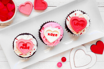 Wall Mural - Fun Valentines Day cupcakes with messages on heart candies. Above view over a white wood background.