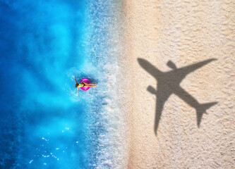 Wall Mural - Aerial view of airplane shadow, swimming beautiful young woman with pink swim ring, tropical sandy beach, sea with waves at summer sunny day. Vacation. Top view of slim girl, azure water, plane shadow