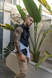Chico joven musculado y tatuado posando con ropa urbana y moderna en zona de bares con palmeras al atardecer
