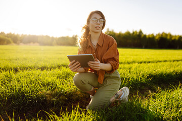 Fototapete - Smart agriculture. A young woman farmer wearing glasses holds a tablet on a green wheat field. A woman agronomist checks the quality of the crop using a digital tablet. Agriculture concept.