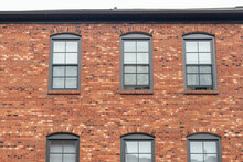 The Exterior Of An Old Red Brick Building With Six Glass Double Hung Windows. The Trim Around The Windows Is Grey. Two Windows Are Open And Four Are Closed. The Roof Is Flat With A Small Vent.