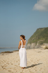 Wall Mural - Back view of beautiful brunette girl in a white linen dress walking by seaside. Travel vacation relaxation abroad lifestyle