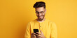 Smiling handsome young man browsing social media on mobile phone while standing on yellow background with lots of copy space on the right