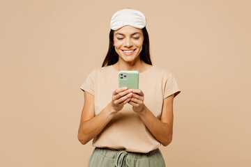 Wall Mural - Young fun calm Latin woman wears pyjamas jam sleep eye mask rest relax at home hold in hand use mobile cell phone isolated on plain pastel light beige background studio. Good mood night nap concept.