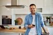 Portrait of an enthusiastic young adult real estate agent holding a cellphone while leaning on a kitchen counter behind him.