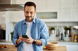 © Jelena - A content young adult man texting his friends on his phone in a kitchen setting.