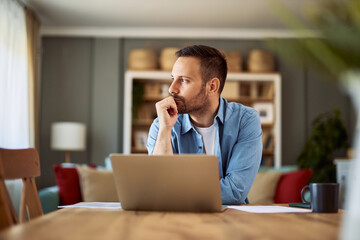 A focused male copywriter looking aside in deep thought with his hand on his chin sitting in front of a laptop.